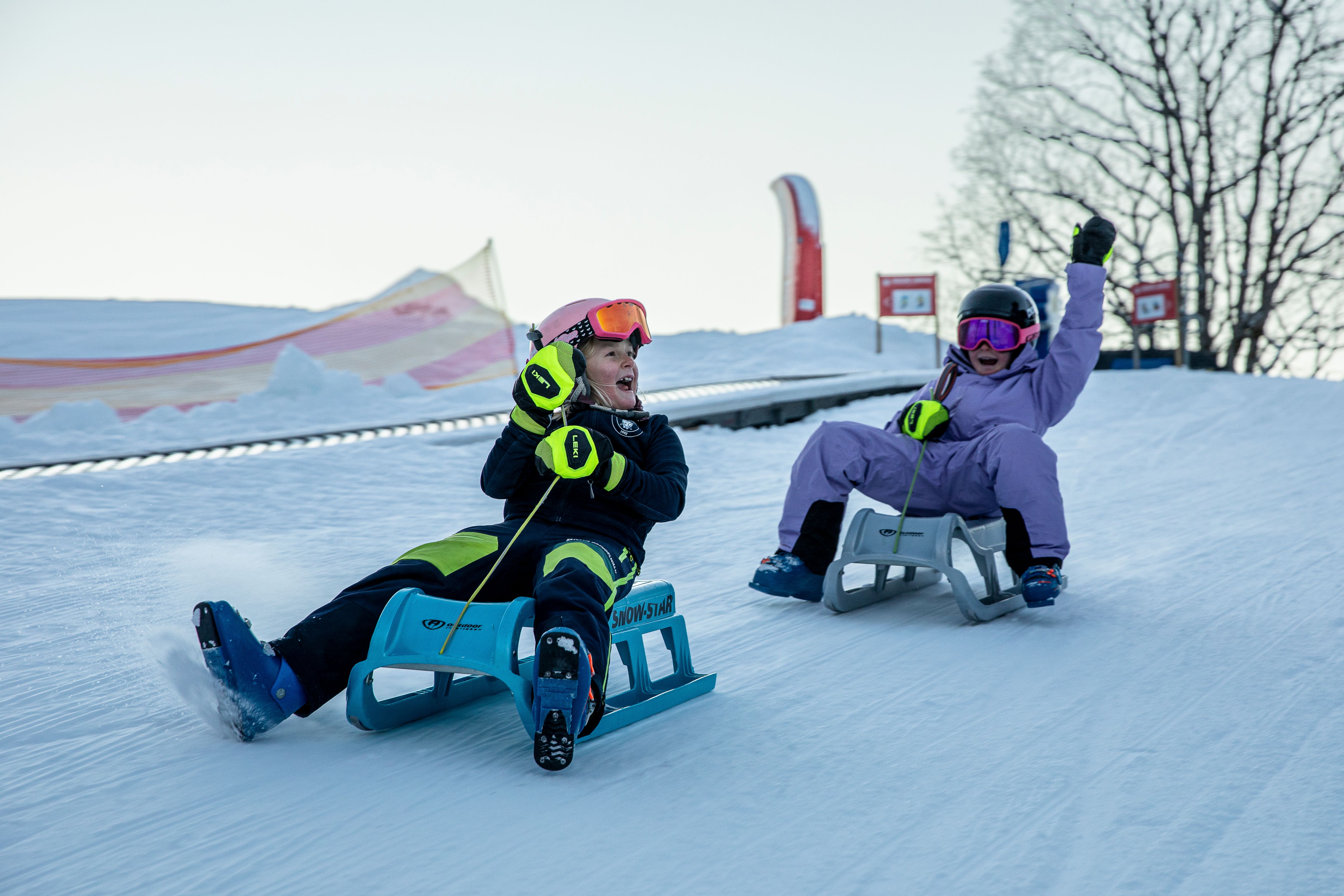Zwei Personen rodeln auf Schnee, tragen Helme und Skibrillen. Hintergrund: verschneite Landschaft.